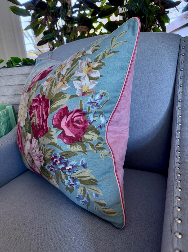 Puede incluir: Una almohada floral con flores rosas y blancas sobre un fondo azul claro. La almohada tiene un borde rosa y est&aacute; sentada en un sof&aacute; azul claro.