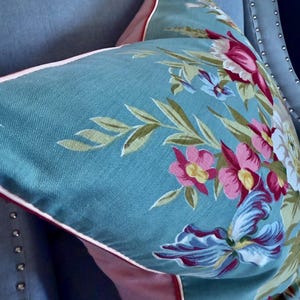 Puede incluir: Una almohada azul turquesa con un patr&oacute;n floral en tonos de rosa, rojo y blanco. La almohada tiene un ribete rosa y blanco.