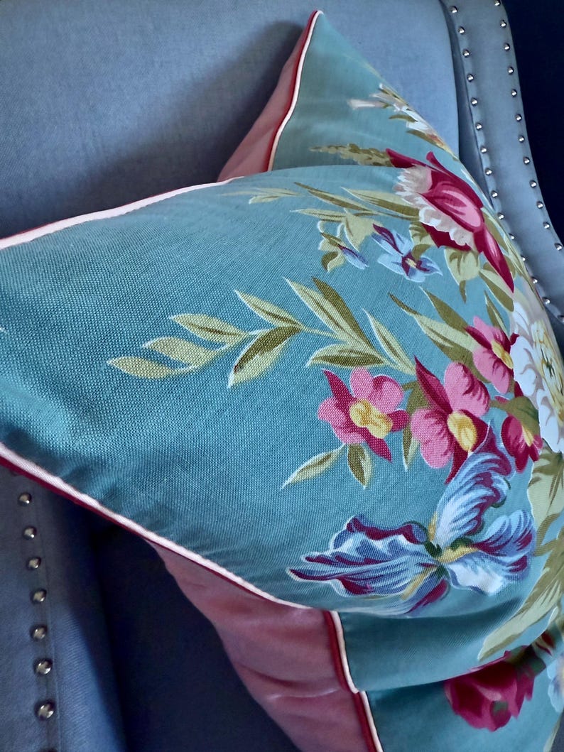 Puede incluir: Una almohada azul turquesa con un patr&oacute;n floral en tonos de rosa, rojo y blanco. La almohada tiene un ribete rosa y blanco.