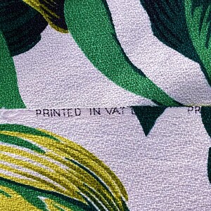 K&ouml;nnte beinhalten: Nahaufnahme eines wei&szlig;en Stoffes mit einem Muster aus gr&uuml;nen und gelben Bl&auml;ttern. Der Stoff hat eine strukturierte Webart und der Text "PRINTED IN VATIC" ist sichtbar.