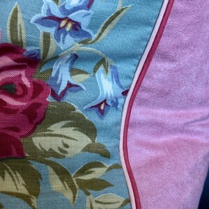 Puede incluir: Una almohada azul y rosa con un patr&oacute;n floral. La almohada tiene una ribetera blanca alrededor del borde y un respaldo de terciopelo rosa.