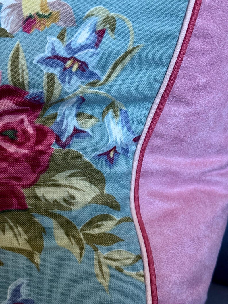 Puede incluir: Una almohada azul y rosa con un patr&oacute;n floral. La almohada tiene una ribetera blanca alrededor del borde y un respaldo de terciopelo rosa.