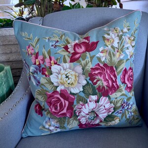 Puede incluir: Una almohada azul floral con ribete blanco se encuentra sobre un sill&oacute;n gris. La almohada presenta un gran patr&oacute;n floral con flores rosas, blancas y verdes.