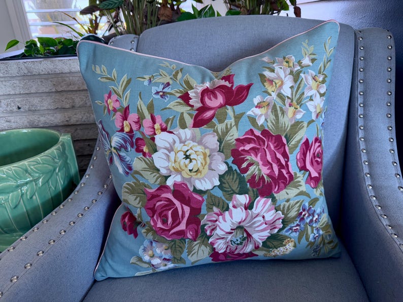 Puede incluir: Una almohada azul floral con ribete blanco se encuentra sobre un sill&oacute;n gris. La almohada presenta un gran patr&oacute;n floral con flores rosas, blancas y verdes.