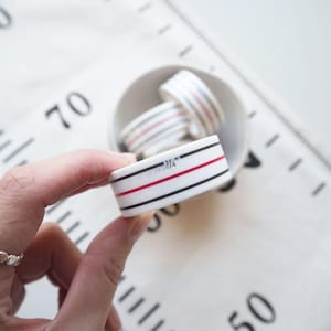 May include: A white ceramic measuring tape with black and red stripes, held in a hand. The tape reads 1/4" and is surrounded by other measuring tapes in a small white bowl. The background features a white fabric with measurement markings.