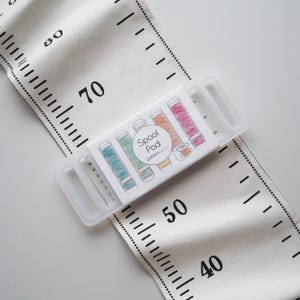 Puede incluir: Una cinta métrica de tela blanca con marcas y números negros, incluyendo 40, 50, 70 y 80. Un recipiente de plástico transparente contiene una tarjeta con la etiqueta "Spool Pod patternface" con ilustraciones de coloridas bobinas de hilo.