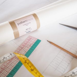 Op de afbeelding: Een rol wit Zweeds traceerpapier met een bruin etiket met de tekst "patterntrace". Een transparante liniaal met rode en groene markeringen, een gele meetlint, een potlood en een schaar liggen op het witte oppervlak.