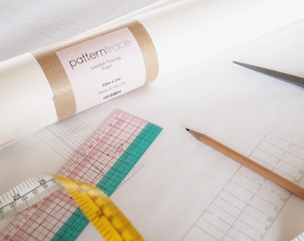 Op de afbeelding: Een rol wit Zweeds traceerpapier met een bruin etiket met de tekst "patterntrace". Een transparante liniaal met rode en groene markeringen, een gele meetlint, een potlood en een schaar liggen op het witte oppervlak.