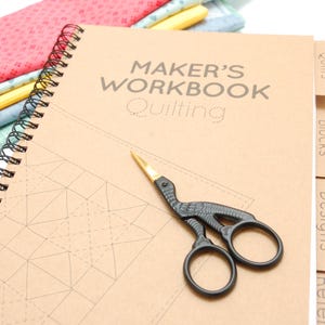 Könnte beinhalten: Ein Spiral gebundenes Maker's Workbook zum Quilten mit dem Titel auf dem Cover. Eine schwarze Storchenschere mit goldenen Klingen liegt auf dem Arbeitsbuch. Bunte Stoffreste sind im Hintergrund zu sehen.