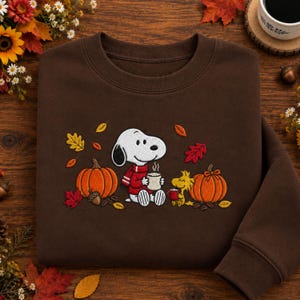 May include: A dark brown crewneck sweatshirt featuring an embroidered design of Snoopy wearing a red scarf and holding a white mug, alongside Woodstock holding a small red mug, surrounded by orange pumpkins and colourful autumn leaves.