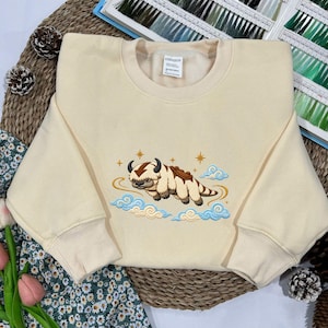 May include: A cream-colored crewneck sweatshirt featuring an embroidered graphic of a furry creature with brown markings and a brown arrow on its head, surrounded by light blue clouds and gold stars. The inner neck label reads M/L 100% Cotton.