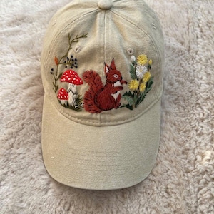 May include: A beige baseball cap with an embroidered design featuring a red squirrel, red mushrooms, and yellow and white flowers. The cap has a curved brim and a button on top. The embroidery is detailed and colorful.