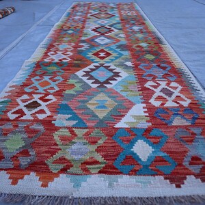 Pode incluir: Um tapete passadeira longo e retangular com um padrão geométrico multicolorido vibrante. O design inclui motivos repetitivos de diamantes e flores estilizadas em tons de vermelho, azul, verde, laranja e branco, com uma borda de franjas.
