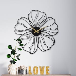 May include: A black metal flower-shaped wall clock with gold-colored hands. The clock is mounted on a white wall above a wooden dresser. The word "LOVE" is spelled out in gold letters on the dresser.