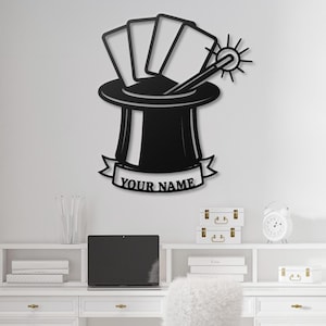 May include: Black metal wall art featuring a magician's hat with playing cards, a wand, and the text "YOUR NAME" on a banner. The design is a silhouette, suitable for home decor.