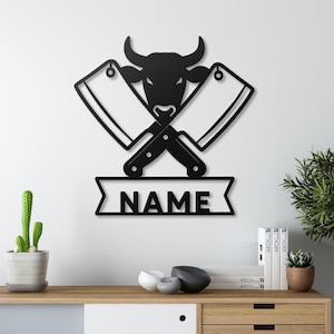 May include: Black metal wall art featuring a bull's head between two crossed cleavers. Below, a banner displays the word "NAME". The artwork is designed for a butcher shop or kitchen decor.