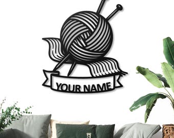 May include: Black metal wall art featuring a yarn ball with two knitting needles and a banner that reads "YOUR NAME". The yarn ball is intricately designed with detailed lines, and the needles are positioned to appear as if they are knitting. This piece is perfect for a craft room.