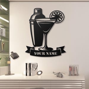 May include: Black metal wall art featuring a cocktail shaker, martini glass with a lemon slice, and a banner with the text "YOUR NAME". The design is a silhouette, suitable for a bar or kitchen.