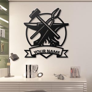 Peut inclure: Décoration murale en métal noir représentant des outils de forgeron : un marteau, des pinces et une enclume, dans un cercle. Une bannière en dessous indique "YOUR NAME". Parfait pour un atelier ou la décoration intérieure.