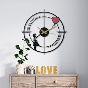 May include: Black metal wall clock featuring a silhouette of a girl reaching for a red heart-shaped balloon. The clock has gold hands and a black face, with a target-like design around the clock face. Decorative wall art.