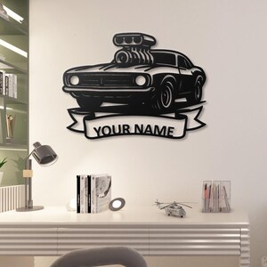 May include: Black metal wall art featuring a classic muscle car design. The car has a large engine and is mounted on a banner that reads "YOUR NAME". This decorative piece is suitable for a garage or man cave.