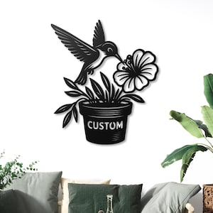 May include: Black metal wall art depicting a hummingbird and flower. The hummingbird is feeding from a flower above a plant pot. The pot has the word "CUSTOM" on it. The artwork is designed for wall mounting.