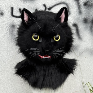 Könnte beinhalten: Eine schwarze Katzenmaske mit gelben Augen, rosa Innenohren und weißen Schnurrhaaren. Die Maske hat eine realistische, pelzige Textur und einen leicht geöffneten Mund, der weiße Zähne und eine rote Zunge zeigt. Der Hintergrund ist eine weiße Wand mit graffitis.