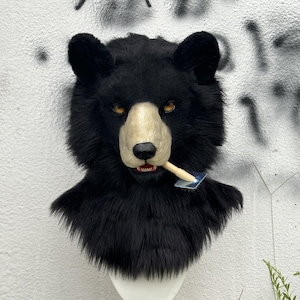 Könnte beinhalten: Eine schwarze Bärenmaske mit hellbrauner Schnauze und gelben Augen. Die Maske hat eine Kunstfellstruktur und einen kleinen Gegenstand im Mund. Die Maske wird vor einer weißen Wand mit grauen Graffiti gezeigt.