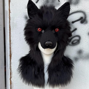 May include: A black wolf mask with red eyes and sharp white teeth. The mask is made of faux fur and has a realistic design. The mask is displayed on a white mannequin head, showcasing the neck and shoulder area.