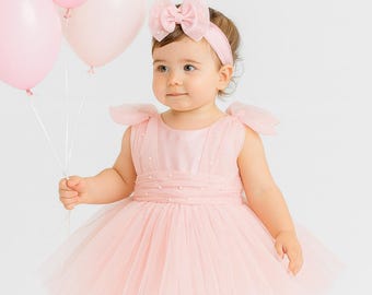 May include: A baby girl in a pink tulle dress and matching headband with a bow, holding pink balloons. The dress features a satin bodice with pearl accents and a layered skirt. The background is plain white.
