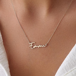 Può includere: Una delicata collana color oro con il nome "Emma" in corsivo. Il ciondolo pende da una sottile catena, appoggiato sulla pelle di una persona. La collana è un gioiello personalizzato.