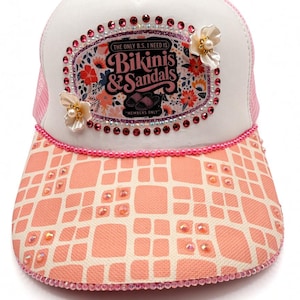 May include: A white and pink trucker hat featuring a central patch with the text "THE ONLY B.S. I NEED IS BIKINIS & SANDALS MEMBERS ONLY" surrounded by pink rhinestones and two white floral embellishments. The brim has a peach grid pattern.