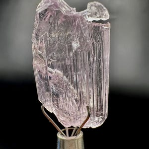 Puede incluir: Un esp&eacute;cimen de cristal de turmalina rosa claro transl&uacute;cido. El cristal est&aacute; montado sobre un soporte met&aacute;lico plateado con pinzas de alambre. El cristal tiene una textura estriada y una forma ligeramente irregular.