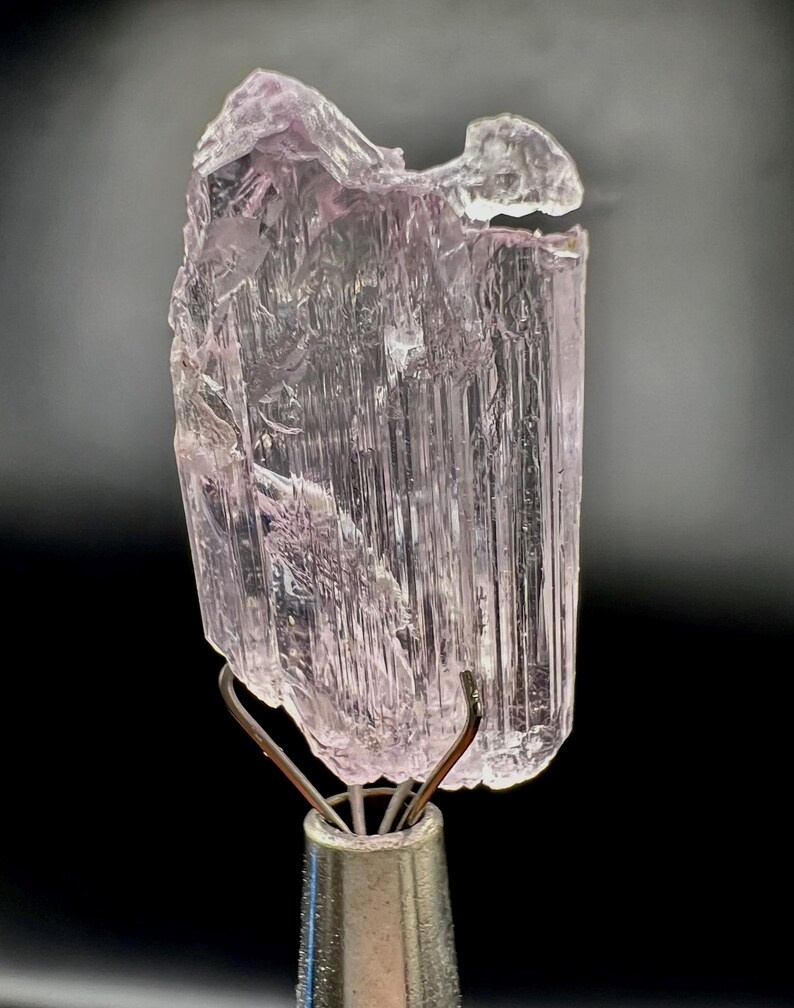 Puede incluir: Un esp&eacute;cimen de cristal de turmalina rosa claro transl&uacute;cido. El cristal est&aacute; montado sobre un soporte met&aacute;lico plateado con pinzas de alambre. El cristal tiene una textura estriada y una forma ligeramente irregular.