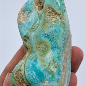 May include: A polished, natural stone with a striking blend of turquoise and brown hues. The stone's surface features swirling patterns and a textured appearance, showcasing its unique geological composition. The stone is held in a hand.