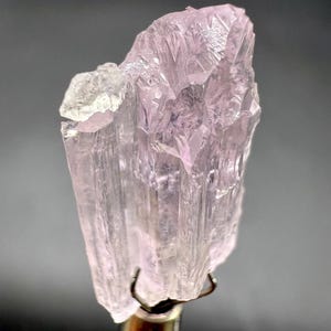 Puede incluir: Un esp&eacute;cimen mineral transl&uacute;cido de color rosa p&aacute;lido con una estructura cristalina. El mineral est&aacute; sujeto por una pinza met&aacute;lica, mostrando su forma natural y tosca. El esp&eacute;cimen tiene un brillo sutil y una gama de tonos rosa p&aacute;lido.