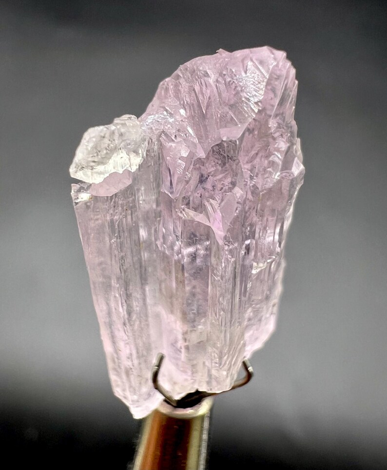 Puede incluir: Un esp&eacute;cimen mineral transl&uacute;cido de color rosa p&aacute;lido con una estructura cristalina. El mineral est&aacute; sujeto por una pinza met&aacute;lica, mostrando su forma natural y tosca. El esp&eacute;cimen tiene un brillo sutil y una gama de tonos rosa p&aacute;lido.