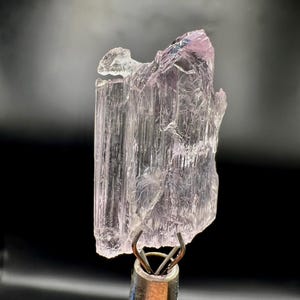 Puede incluir: Un esp&eacute;cimen de cristal transl&uacute;cido de color rosa p&aacute;lido. El cristal est&aacute; sujeto por una pinza met&aacute;lica, sobre un fondo negro. El cristal tiene un aspecto texturizado y en capas con un tono rosa ligeramente m&aacute;s oscuro en la parte superior.