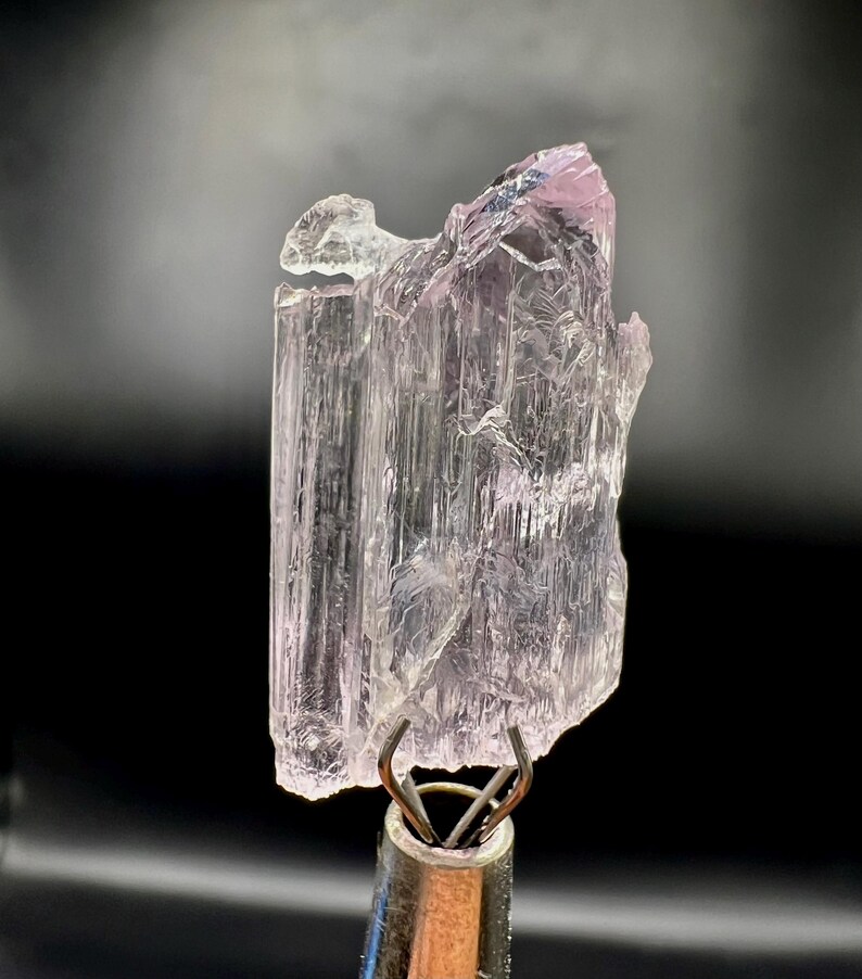 Puede incluir: Un esp&eacute;cimen de cristal transl&uacute;cido de color rosa p&aacute;lido. El cristal est&aacute; sujeto por una pinza met&aacute;lica, sobre un fondo negro. El cristal tiene un aspecto texturizado y en capas con un tono rosa ligeramente m&aacute;s oscuro en la parte superior.
