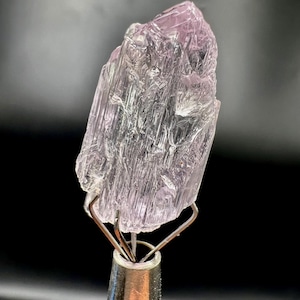Puede incluir: Un esp&eacute;cimen de cristal transl&uacute;cido de color p&uacute;rpura claro, con una textura estriada. El cristal est&aacute; montado sobre un soporte met&aacute;lico plateado, sobre un fondo oscuro. El color del cristal var&iacute;a del lavanda p&aacute;lido al violeta m&aacute;s intenso.