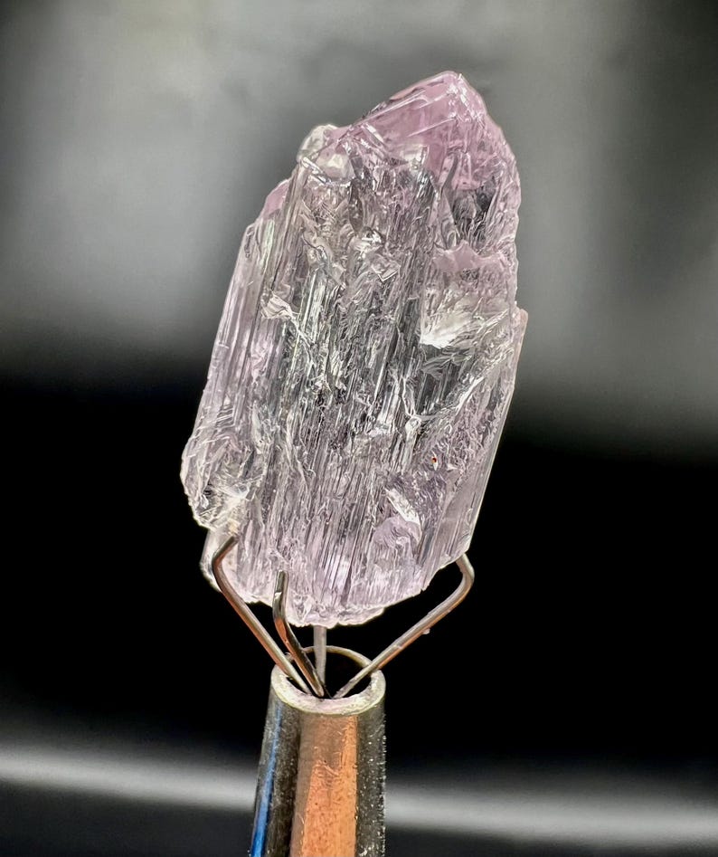 Puede incluir: Un esp&eacute;cimen de cristal transl&uacute;cido de color p&uacute;rpura claro, con una textura estriada. El cristal est&aacute; montado sobre un soporte met&aacute;lico plateado, sobre un fondo oscuro. El color del cristal var&iacute;a del lavanda p&aacute;lido al violeta m&aacute;s intenso.