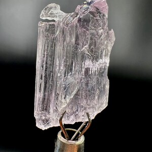 Puede incluir: Un esp&eacute;cimen de cristal transl&uacute;cido, de color p&uacute;rpura claro, con un aspecto texturizado y en capas. El cristal est&aacute; sujeto por una pinza met&aacute;lica sobre un fondo negro. La parte superior del cristal tiene un tono p&uacute;rpura m&aacute;s oscuro.