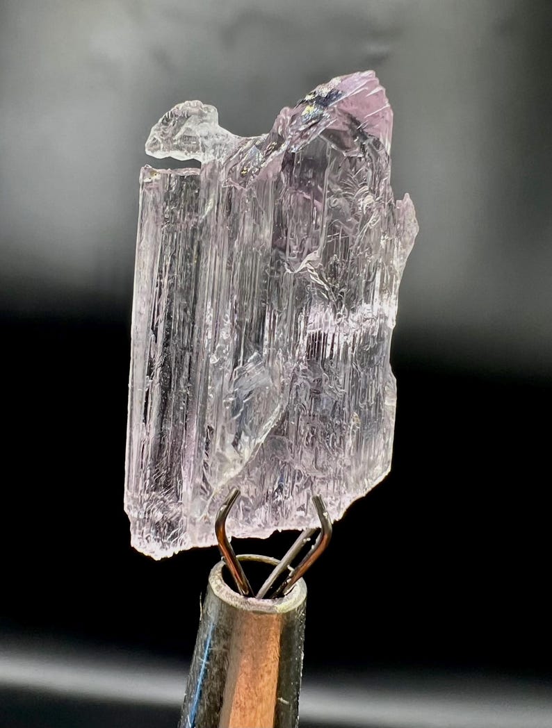 Puede incluir: Un esp&eacute;cimen de cristal transl&uacute;cido, de color p&uacute;rpura claro, con un aspecto texturizado y en capas. El cristal est&aacute; sujeto por una pinza met&aacute;lica sobre un fondo negro. La parte superior del cristal tiene un tono p&uacute;rpura m&aacute;s oscuro.