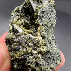 May include: A close-up of a mineral specimen featuring a cluster of dark green, needle-like crystals. The crystals are set against a matrix of lighter green and gray, creating a textured appearance. The mineral is a natural, raw stone.