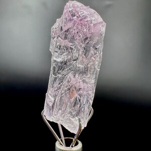 Puede incluir: Un esp&eacute;cimen de cristal rosa claro transl&uacute;cido con una superficie rugosa y texturizada. El cristal est&aacute; montado sobre un soporte met&aacute;lico plateado sobre un fondo negro. El color del cristal var&iacute;a de transparente a rosa suave.