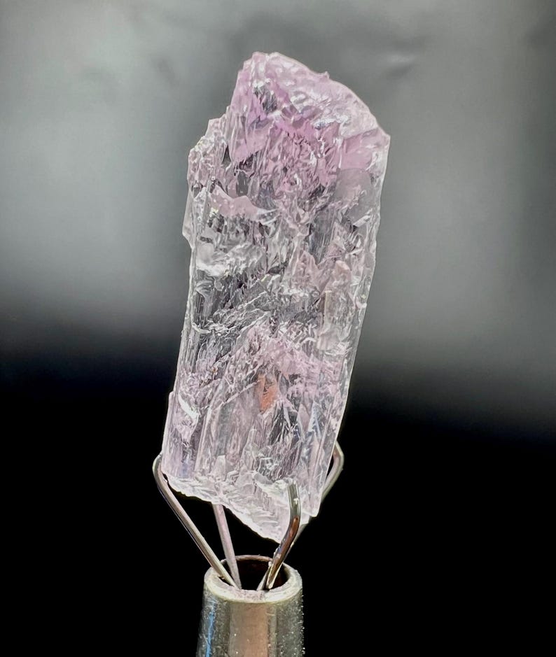 Puede incluir: Un esp&eacute;cimen de cristal rosa claro transl&uacute;cido con una superficie rugosa y texturizada. El cristal est&aacute; montado sobre un soporte met&aacute;lico plateado sobre un fondo negro. El color del cristal var&iacute;a de transparente a rosa suave.