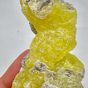 Puede incluir: Un vibrante espécimen mineral de azufre amarillo con una superficie rugosa y texturizada. El mineral tiene parches de inclusiones grises y negras, creando un contraste llamativo. El espécimen se sostiene en una mano, mostrando su forma natural.
