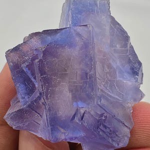 May include: A close-up of a fluorite crystal cluster. The crystal has a layered, geometric structure with a gradient of colors, transitioning from deep purple to a lighter blue. The surface has a crystalline texture, reflecting light.