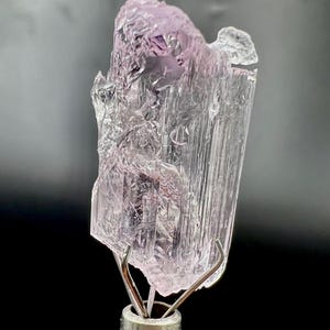 Puede incluir: Un esp&eacute;cimen de cristal transl&uacute;cido de color p&uacute;rpura claro con una estructura cristalina. El cristal est&aacute; montado sobre un soporte met&aacute;lico plateado. El fondo es un degradado de gris y negro.