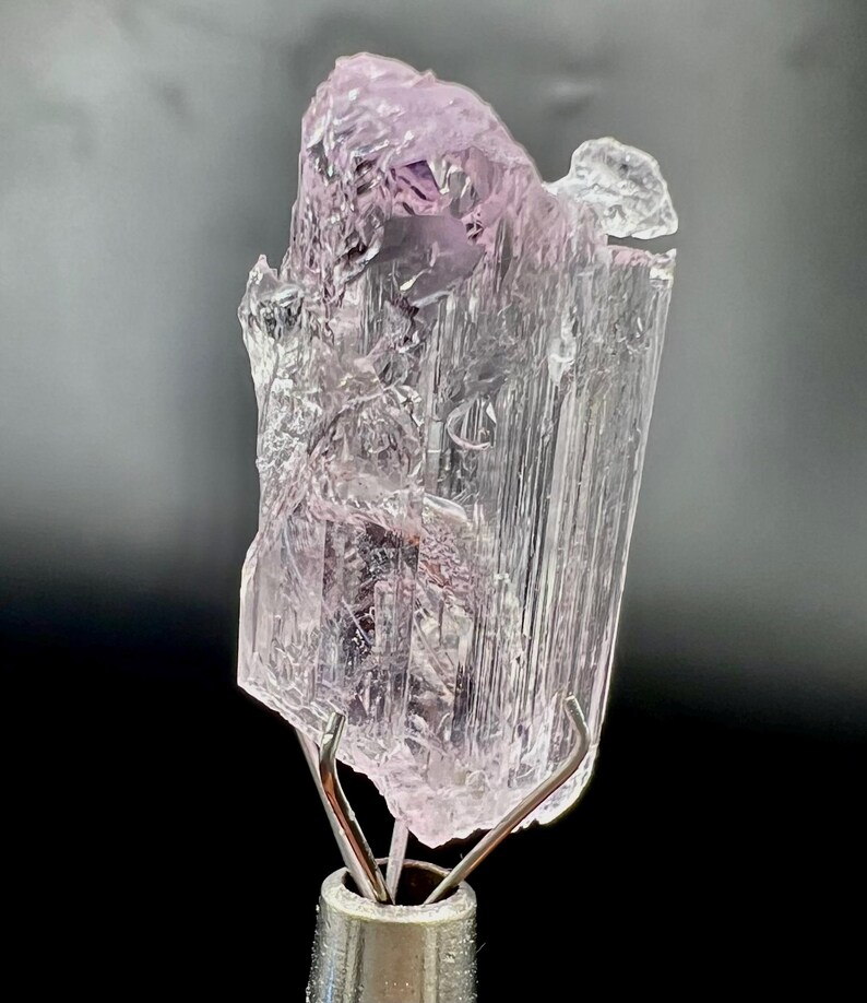 Puede incluir: Un esp&eacute;cimen de cristal transl&uacute;cido de color p&uacute;rpura claro con una estructura cristalina. El cristal est&aacute; montado sobre un soporte met&aacute;lico plateado. El fondo es un degradado de gris y negro.